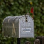 A traditional U.S. mailbox with red flag against a lush green blurred background.